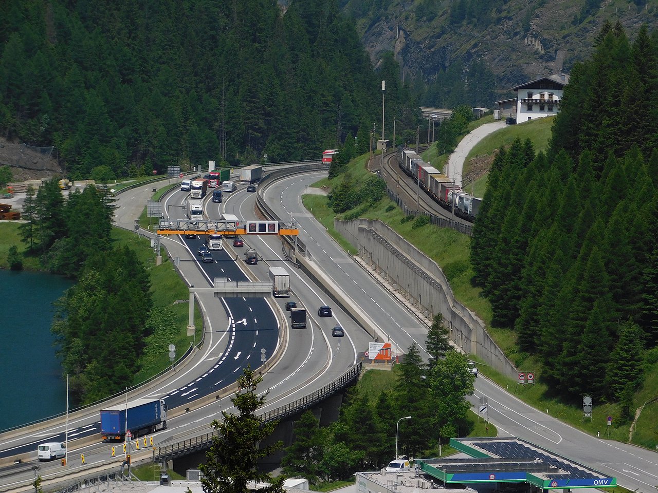 image showing the Brenner Highway and the Brenner railway side by side at Venntal, close to Brenner lake and the highest point of Brenner pass. There are cars and trucks on the Brenner Highway and a freight train on the Brenner railway